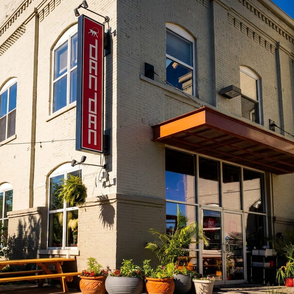 Exterior of Dan Dan restaurant, a light brick building with large windows, a red sign, and outdoor seating.