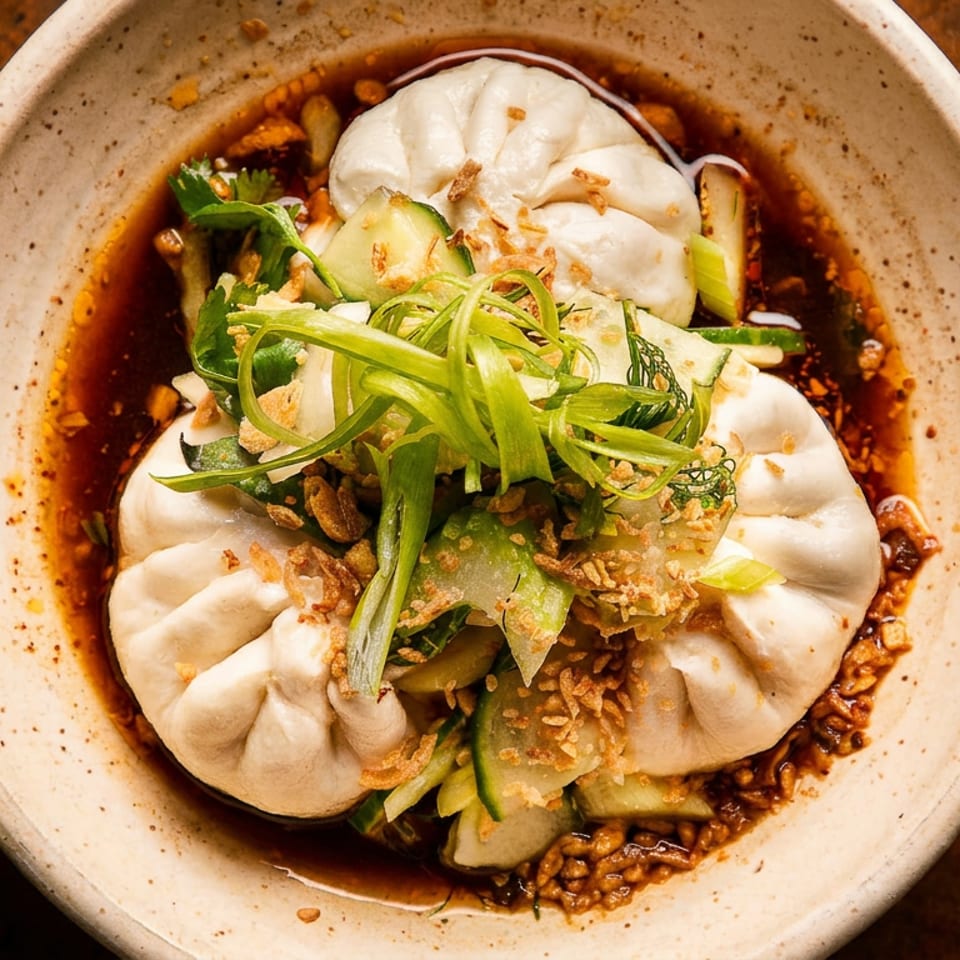 Three steamed buns in a bowl of sauce, topped with sliced cucumbers, green onions, and fried garlic.